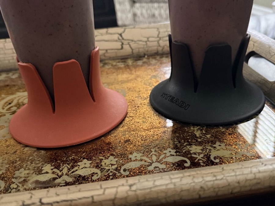 Two drink bases — coral and black — each holding a tall glass of purple berry smoothie, standing on an ornate gold-patterned serving tray in a living room with a patterned armchair behind